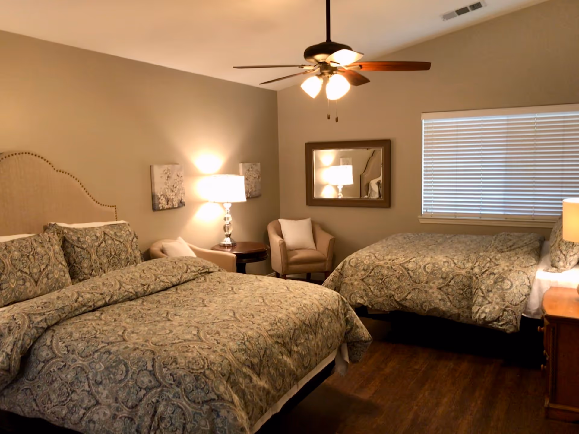 A cozy bedroom with two beds covered in patterned bedspreads, a ceiling fan with lights, a window with blinds, a small round table with a lamp, two armchairs, a wall mirror, and wooden flooring.
