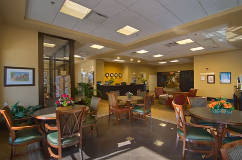 A well-lit common area in a senior living facility with multiple round tables and chairs arranged for seating. The room features a reception desk in the background, decorative plants, framed artwork on the walls, and a cozy seating area with armchairs. The ceiling has recessed lighting and the floor is tiled.