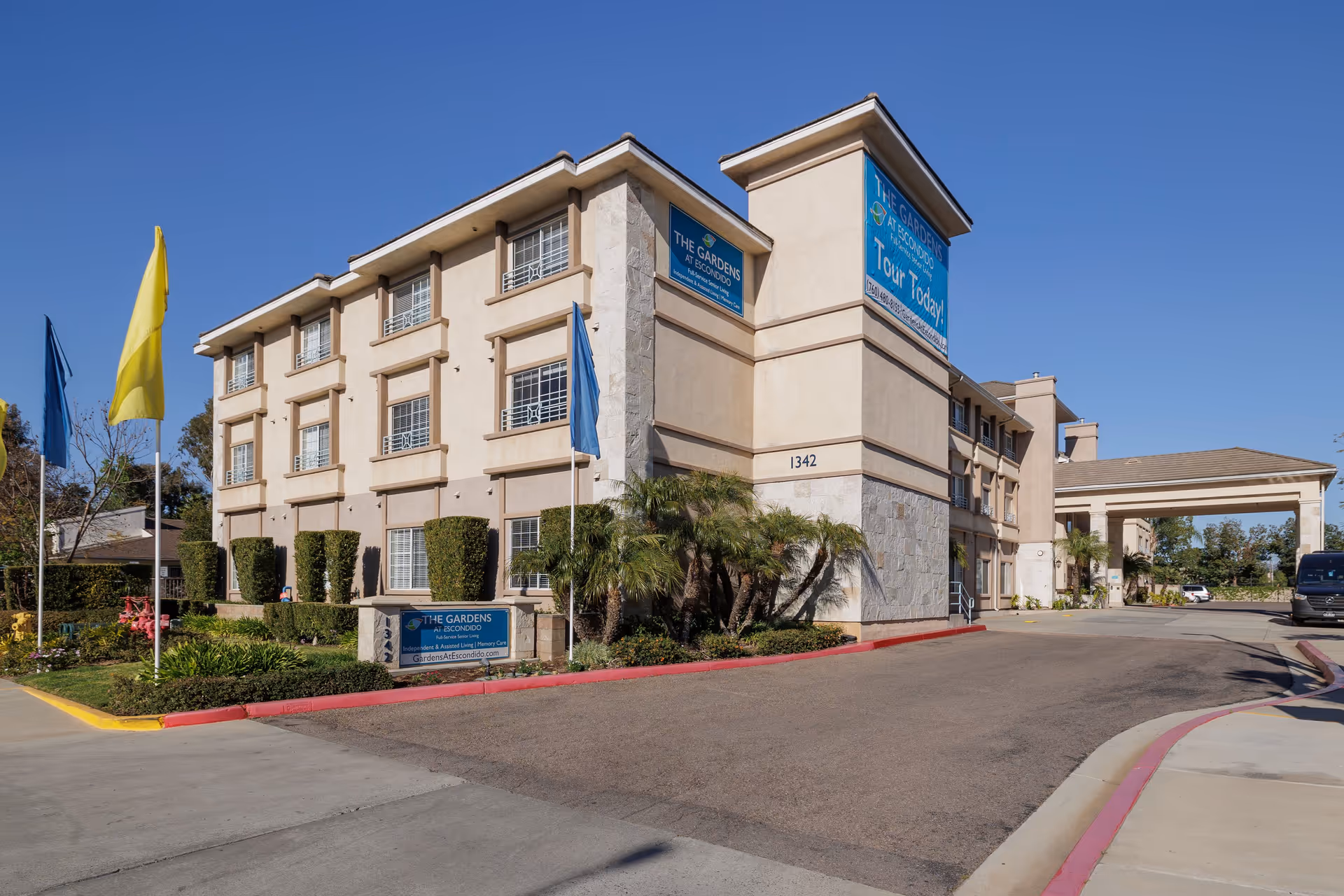 Exterior view of The Gardens at Escondido senior living facility building with beige walls, multiple windows, and a covered entrance. There are flags on poles and well-maintained landscaping with bushes and palm trees. The sky is clear and blue.