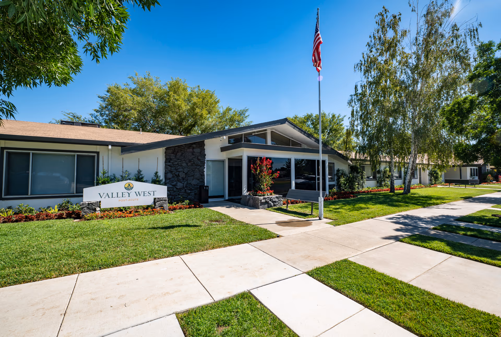 Exterior view of Valley West Post-Acute facility on a sunny day, showing a single-story building with a stone accent wall, large windows, a flagpole with an American flag, well-maintained green lawn, trees, and a concrete walkway leading to the entrance.
