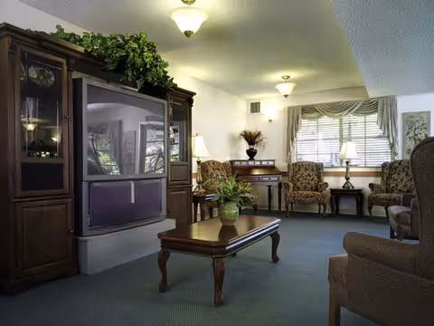 A cozy living room with a large wooden entertainment center holding a TV, a wooden coffee table with a potted plant, several upholstered armchairs, a window with drapes, and soft lighting from ceiling fixtures and lamps.