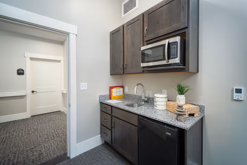 A small kitchenette area with dark wood cabinets, a granite countertop, a stainless steel microwave mounted above the counter, a small sink, and a mini refrigerator below. On the countertop, there are decorative items including a small plant, stacked containers, and a woven basket. The kitchenette is adjacent to a hallway with a door labeled 219.