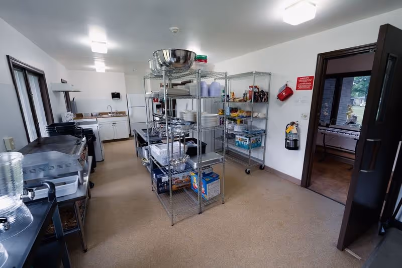 A clean and organized kitchen area with metal shelving units holding various kitchen supplies, dishes, and food items. There is a countertop with a sink and cabinets along the back wall, and a large window on the left side. The room has beige flooring and white walls, with a fire extinguisher and a red fire safety sign mounted on the wall near a doorway leading to another room.