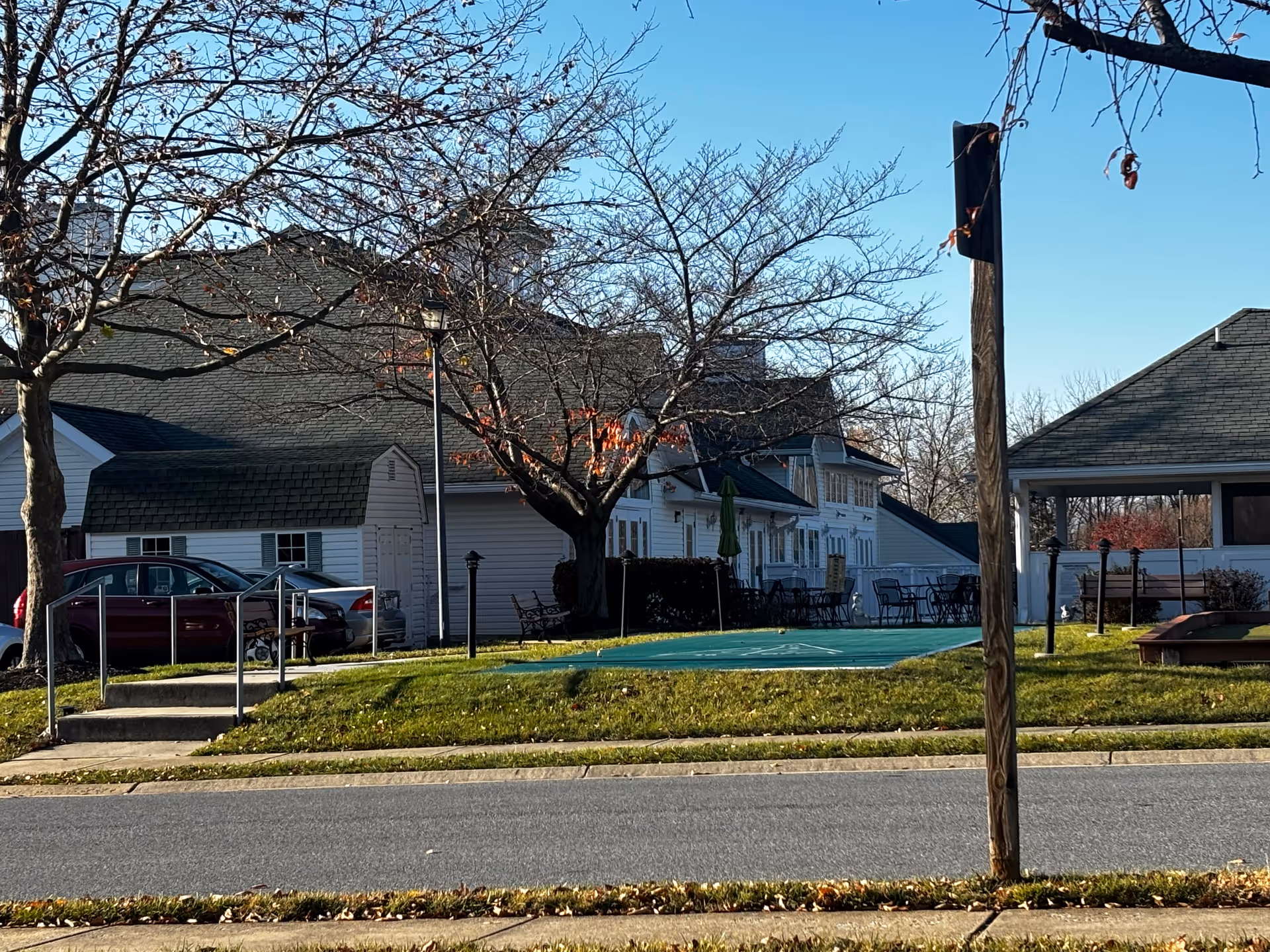Outdoor view of a senior living facility with a green shuffleboard court, patio tables and chairs, leafless trees, and a building with white siding and dark roof under a clear blue sky.