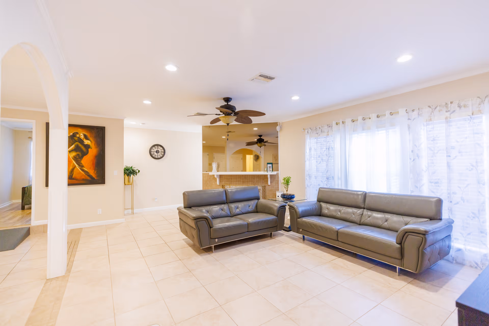 A bright living room with two gray leather sofas facing each other, beige tiled floor, ceiling fans with lights, large windows with sheer white curtains, a wall clock, a painting of a dancing couple, and a small plant on a stand.