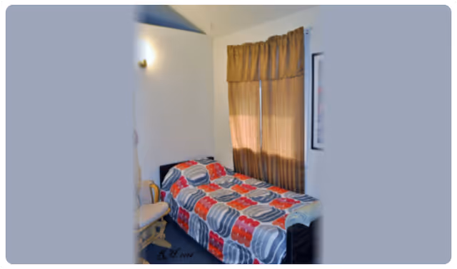 A small bedroom with a single bed covered in a colorful quilt featuring red, gray, and white patterns. There is a wooden rocking chair next to the bed and a window with brown curtains letting in some light. The walls are light-colored, and a wall-mounted light fixture is visible above the bed.