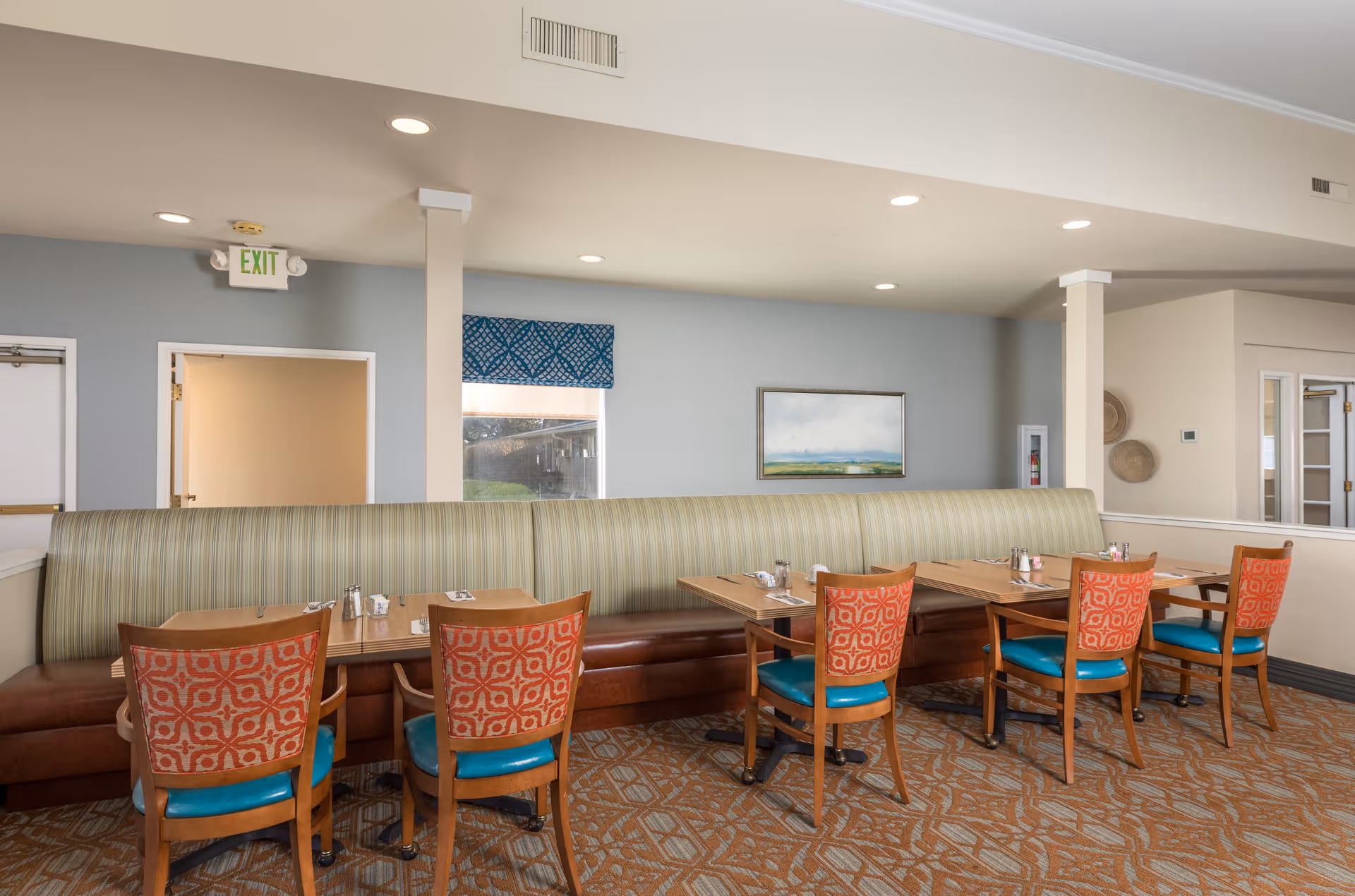 A dining room with a long striped banquette, wooden tables and patterned chairs under recessed lighting.