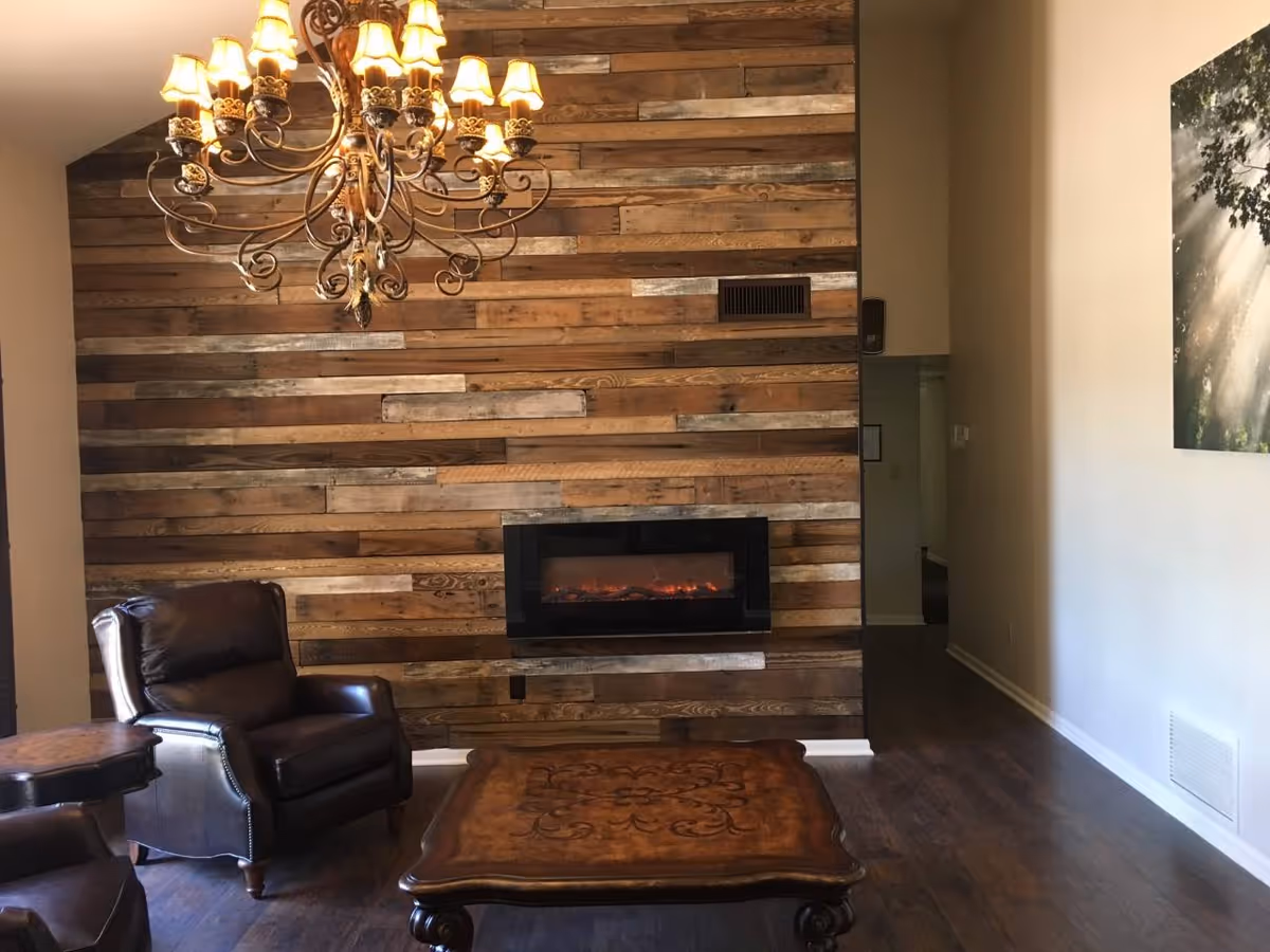 Cozy living room with a wood-plank accent wall, wall-mounted electric fireplace, leather armchairs, an ornate chandelier, and a wooden coffee table.