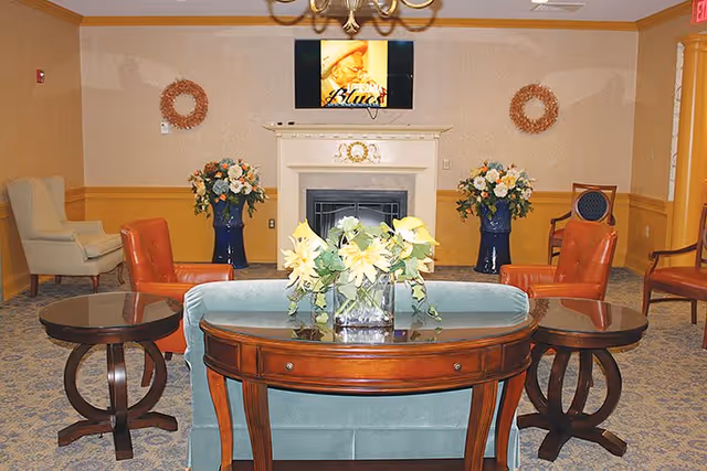 A cozy living room area with a light blue sofa facing a white fireplace. On the sofa's back is a wooden console table with a glass vase holding yellow flowers. Two round wooden side tables flank the sofa. There are two orange armchairs and a beige armchair arranged around the room. Two large blue vases with flower arrangements are placed symmetrically on either side of the fireplace. A flat-screen TV is mounted above the fireplace displaying an image of a woman. The walls are decorated with two circular wreaths and the room has a patterned carpet and warm lighting.