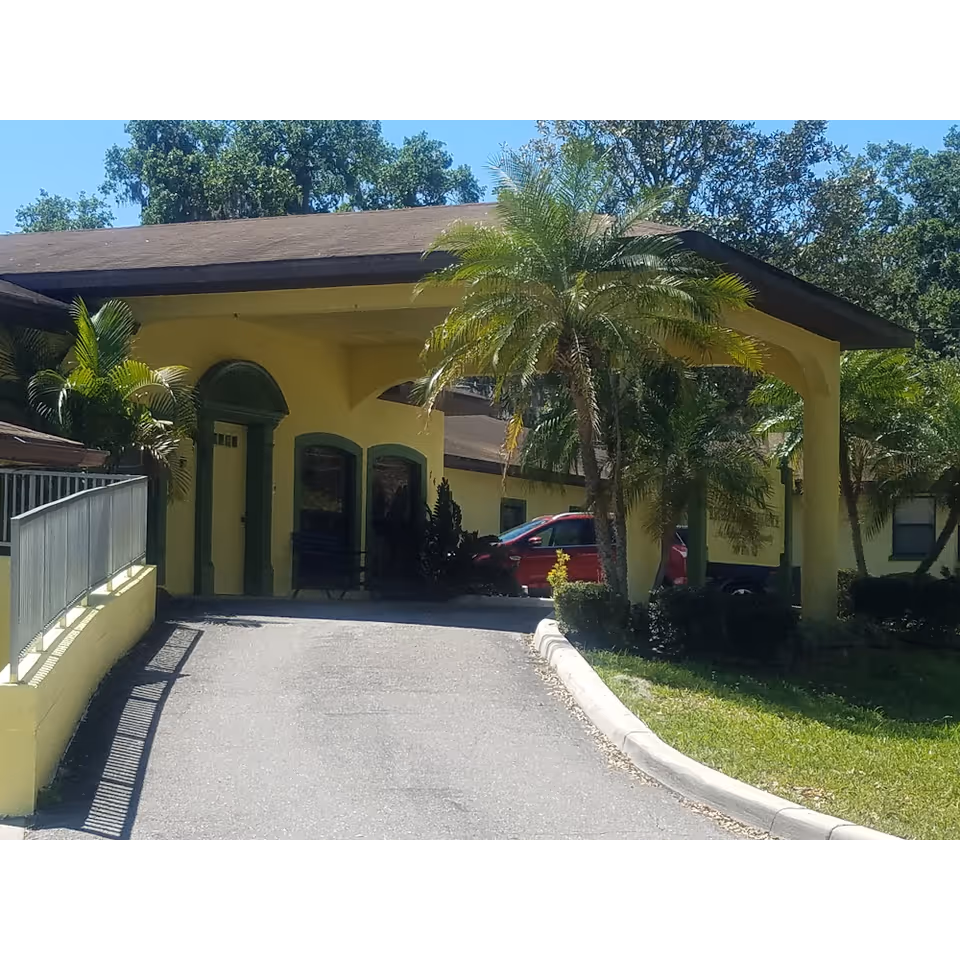 Exterior view of Lakeland Manor Assisted Living Facility showing a yellow building with green trim, a covered driveway entrance, palm trees, and a red car parked near the entrance.
