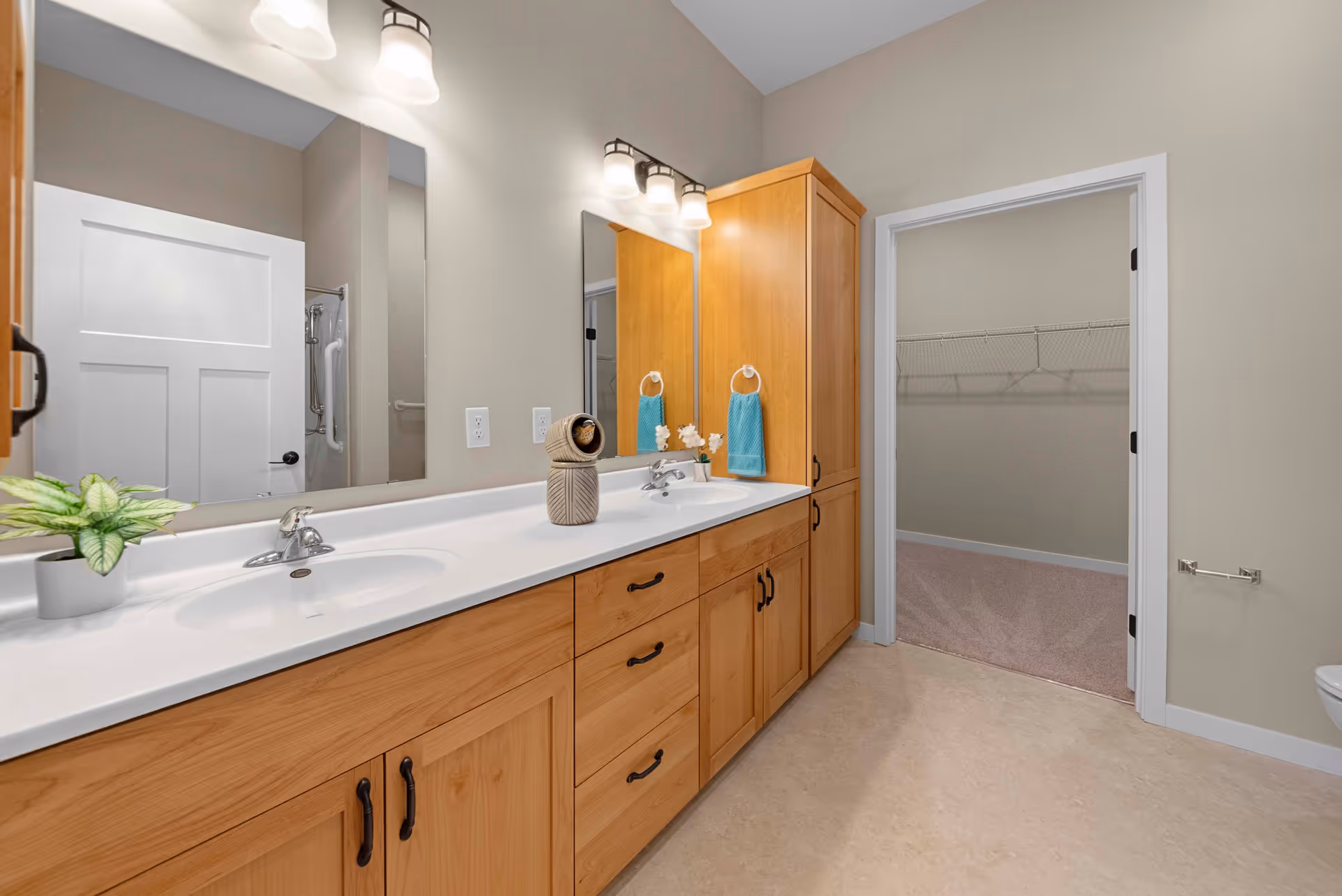 A bathroom interior featuring a double sink vanity with wooden cabinets and two mirrors above. The countertop has a small potted plant and decorative items. There are two light fixtures above the mirrors. A door on the right side leads to a walk-in closet with wire shelving. The bathroom has beige walls and a light-colored floor.