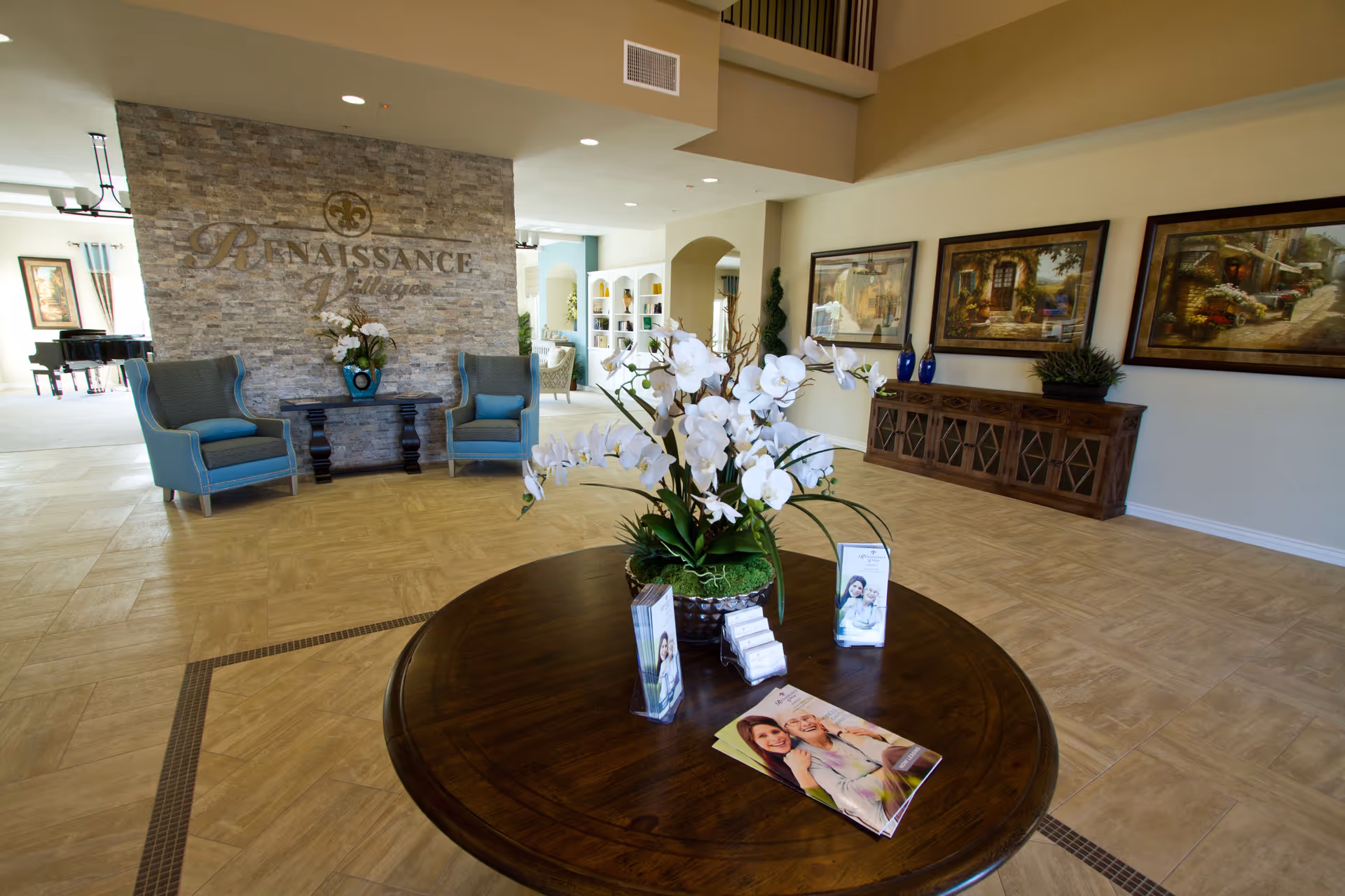 Spacious and well-lit interior common area of Renaissance Village Murrieta featuring a round wooden table with a floral arrangement and brochures, two blue armchairs flanking a stone accent wall with the Renaissance Village logo, framed artwork on the walls, and a glimpse of additional seating and a piano in the background.