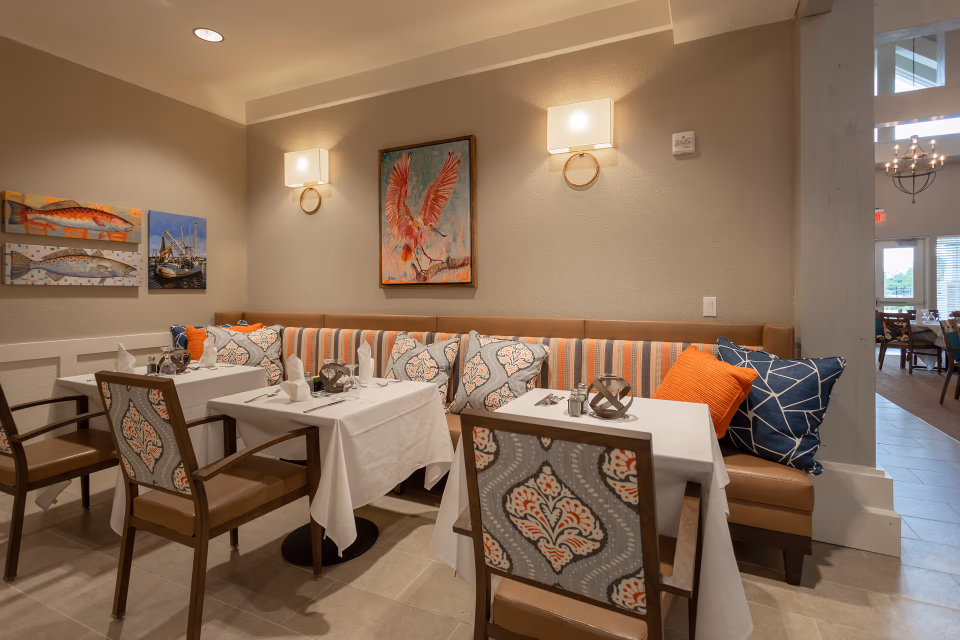 A cozy dining area with two tables covered in white tablecloths, each set with napkins, silverware, and decorative items. The seating includes a cushioned bench with striped and patterned pillows, and wooden chairs with patterned upholstery. The walls are decorated with paintings of fish and a bird, and two wall-mounted lights provide warm illumination.