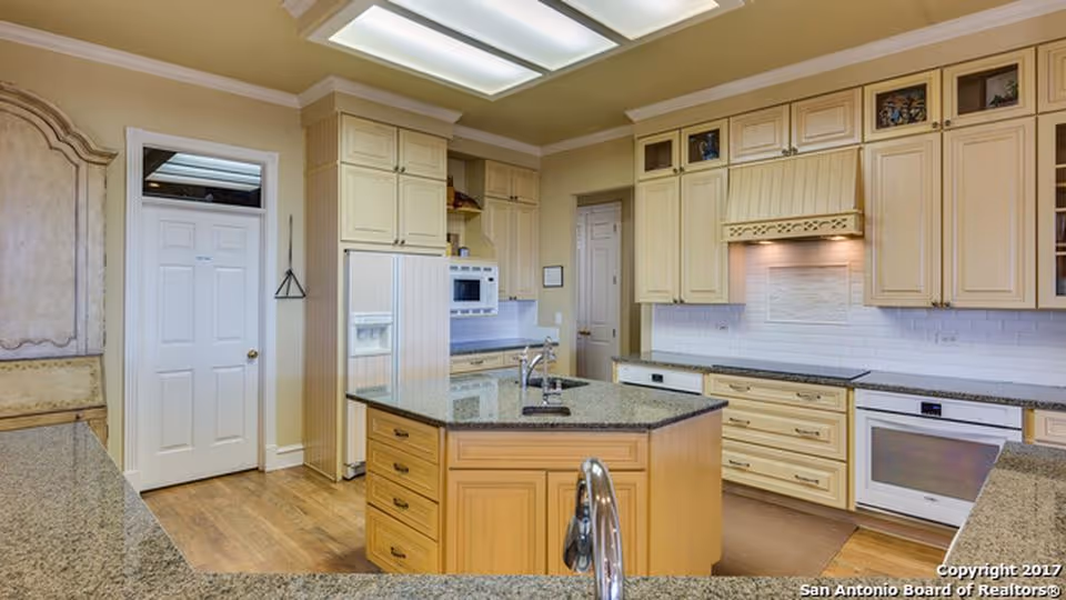 A spacious kitchen with light wood cabinetry, granite countertops, a central island with a sink, built-in microwave, oven, refrigerator, and a white subway tile backsplash. The floor is wooden, and there is a door leading to another room.