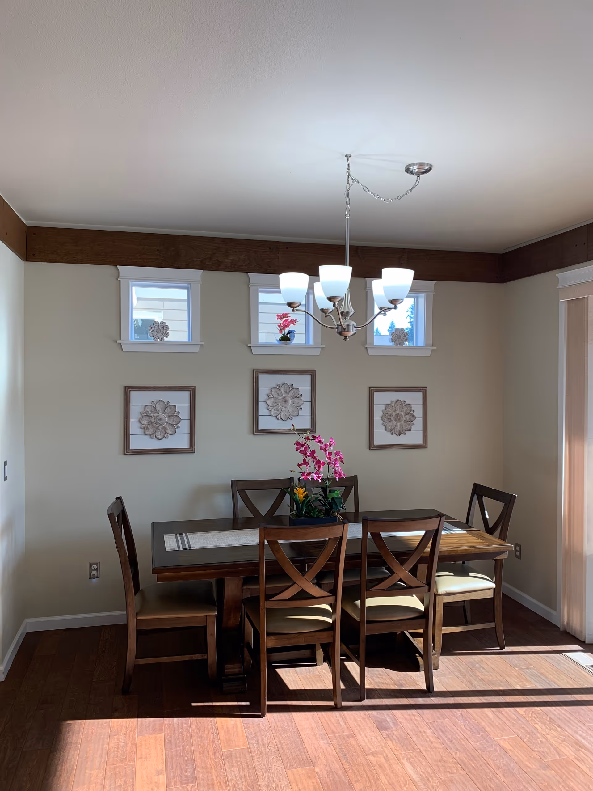 Sunlit dining room with a wooden table and six chairs, a chandelier overhead, a floral centerpiece, and framed wall art beneath small windows.