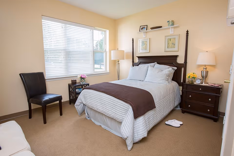 Sunlit bedroom with a four-poster bed, nightstands, a chair, and a window with blinds.