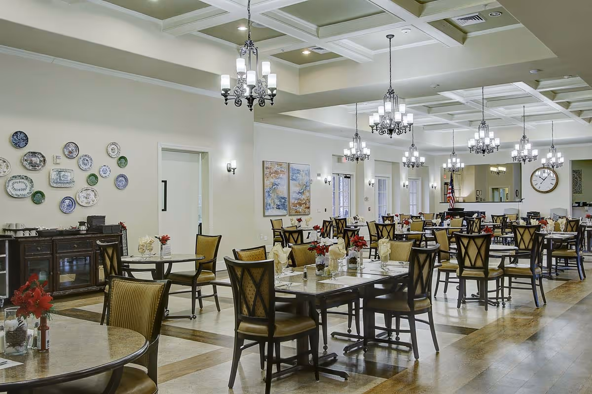 A spacious dining room with multiple tables and chairs arranged neatly. Each table is set with folded napkins, glassware, and small floral centerpieces. The room features elegant chandeliers hanging from a coffered ceiling, light-colored walls adorned with decorative plates and paintings, and large windows allowing natural light to enter.