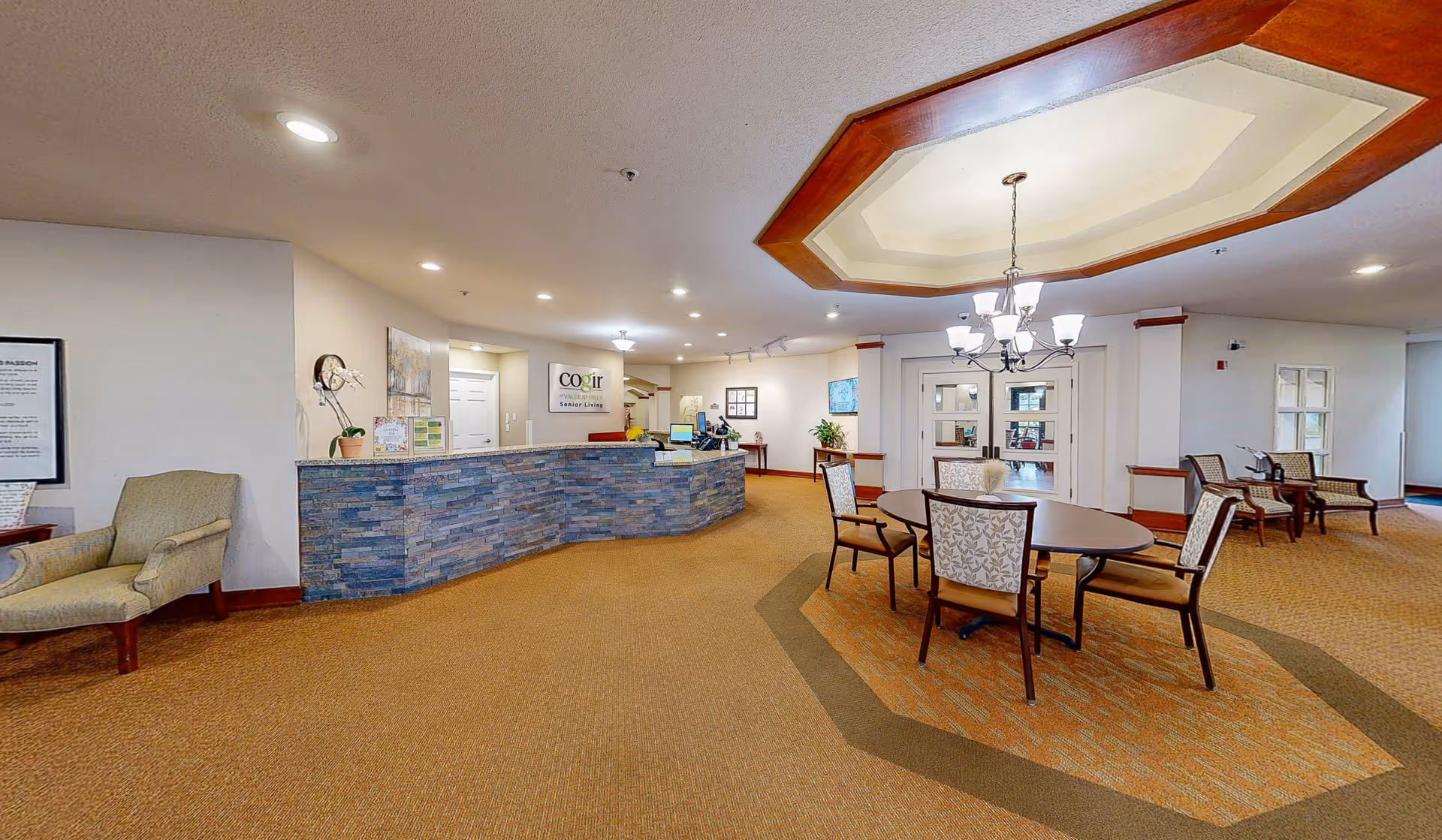 Interior view of a senior living facility lobby with a curved stone reception desk, a round table with four chairs in the center, additional seating chairs along the walls, and a decorative ceiling light fixture.