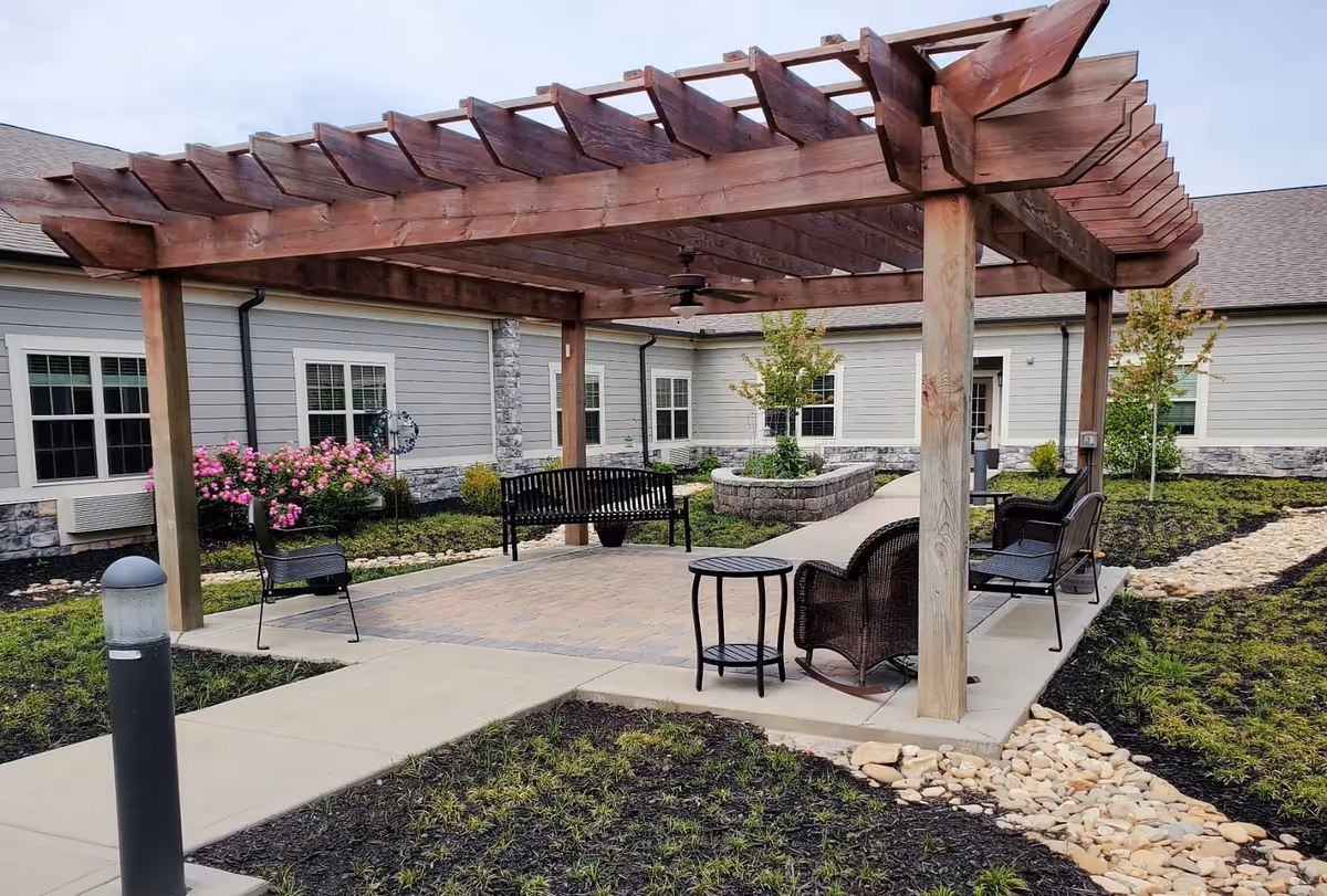 Outdoor seating area with a wooden pergola, black metal benches, a wicker chair, and a small table on a paved patio surrounded by landscaped garden beds with flowers and small trees, adjacent to a light gray building with white-trimmed windows.