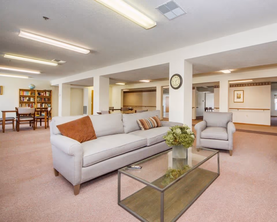 A spacious living room area in Regency Heights featuring a light gray sofa with two pillows, a matching armchair, and a glass coffee table with a vase of green flowers. In the background, there is a wooden bookshelf filled with books and a table with chairs. The room has beige carpeting, white walls, and fluorescent ceiling lights.