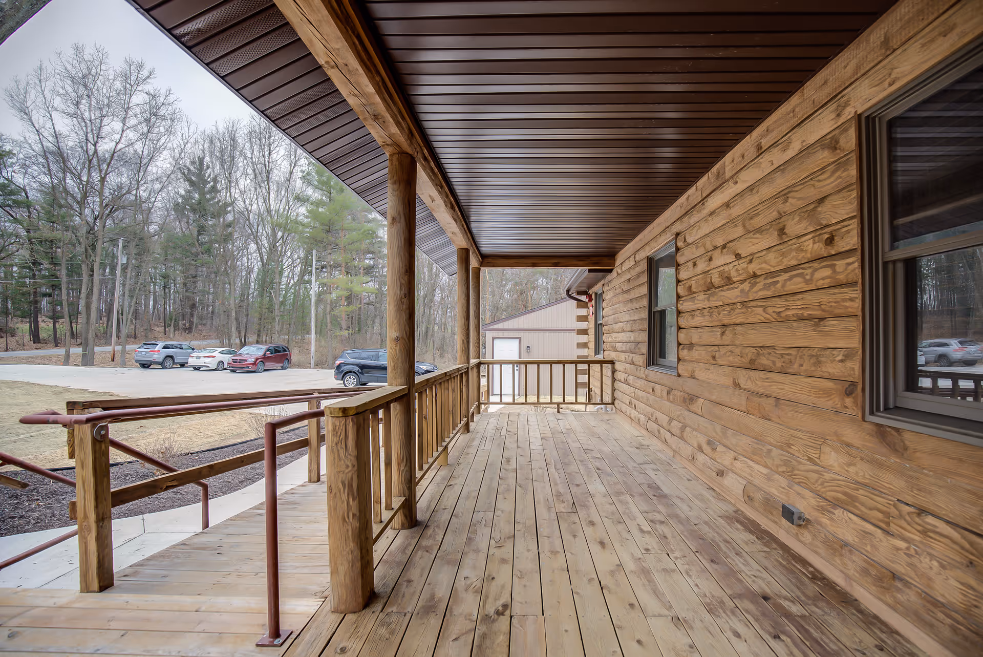 Covered wooden porch with railing and ramp leading down to a parking area with several cars. The porch is attached to a building with wooden siding and two windows. Trees and a small building are visible in the background.