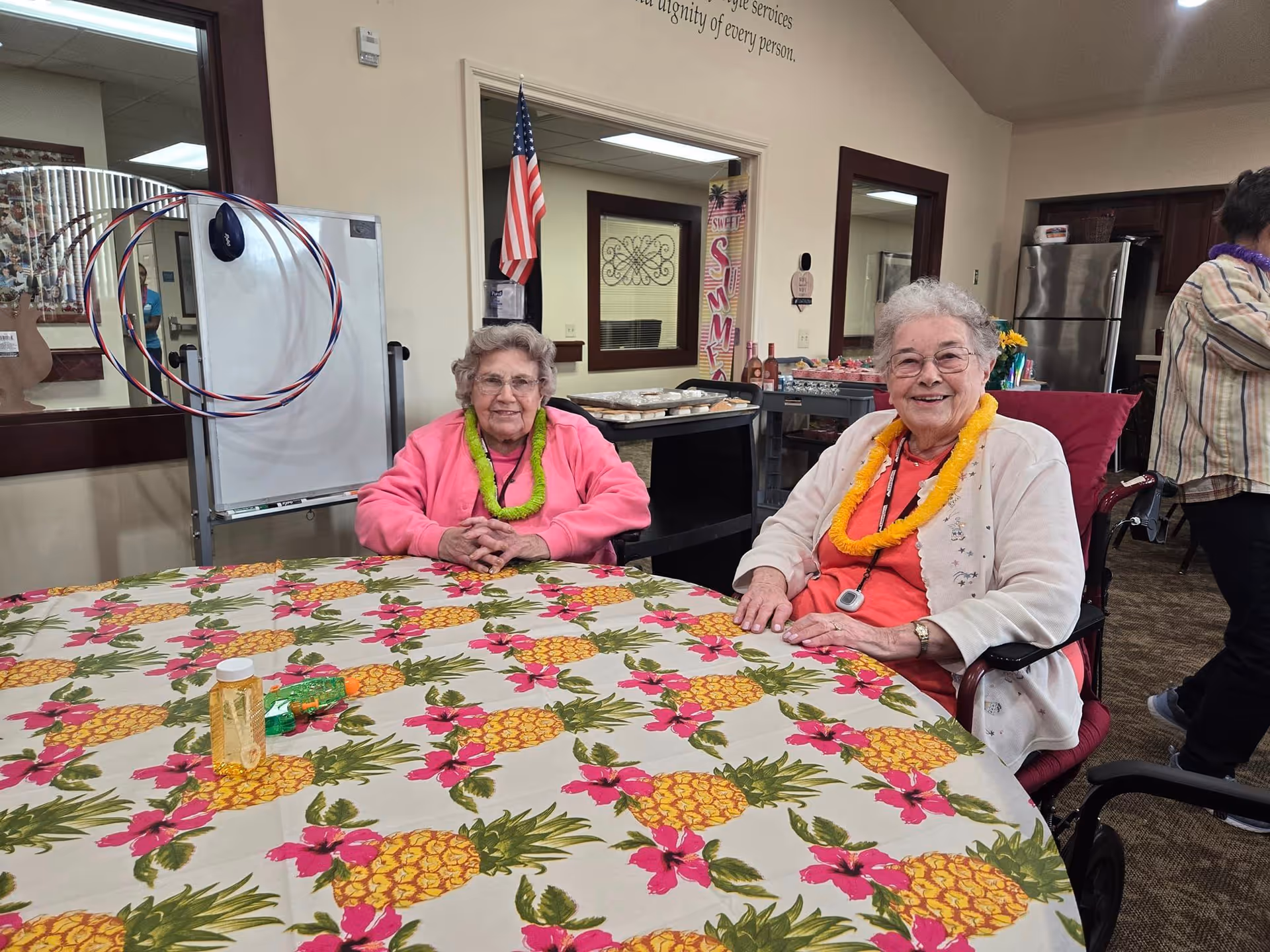 Two elderly women sitting at a round table covered with a pineapple and pink flower patterned tablecloth in a common room. Both women are wearing colorful leis and smiling. Behind them is a whiteboard with hula hoops hanging on it, an American flag, and a kitchen area with a refrigerator. Another person is partially visible standing to the right.
