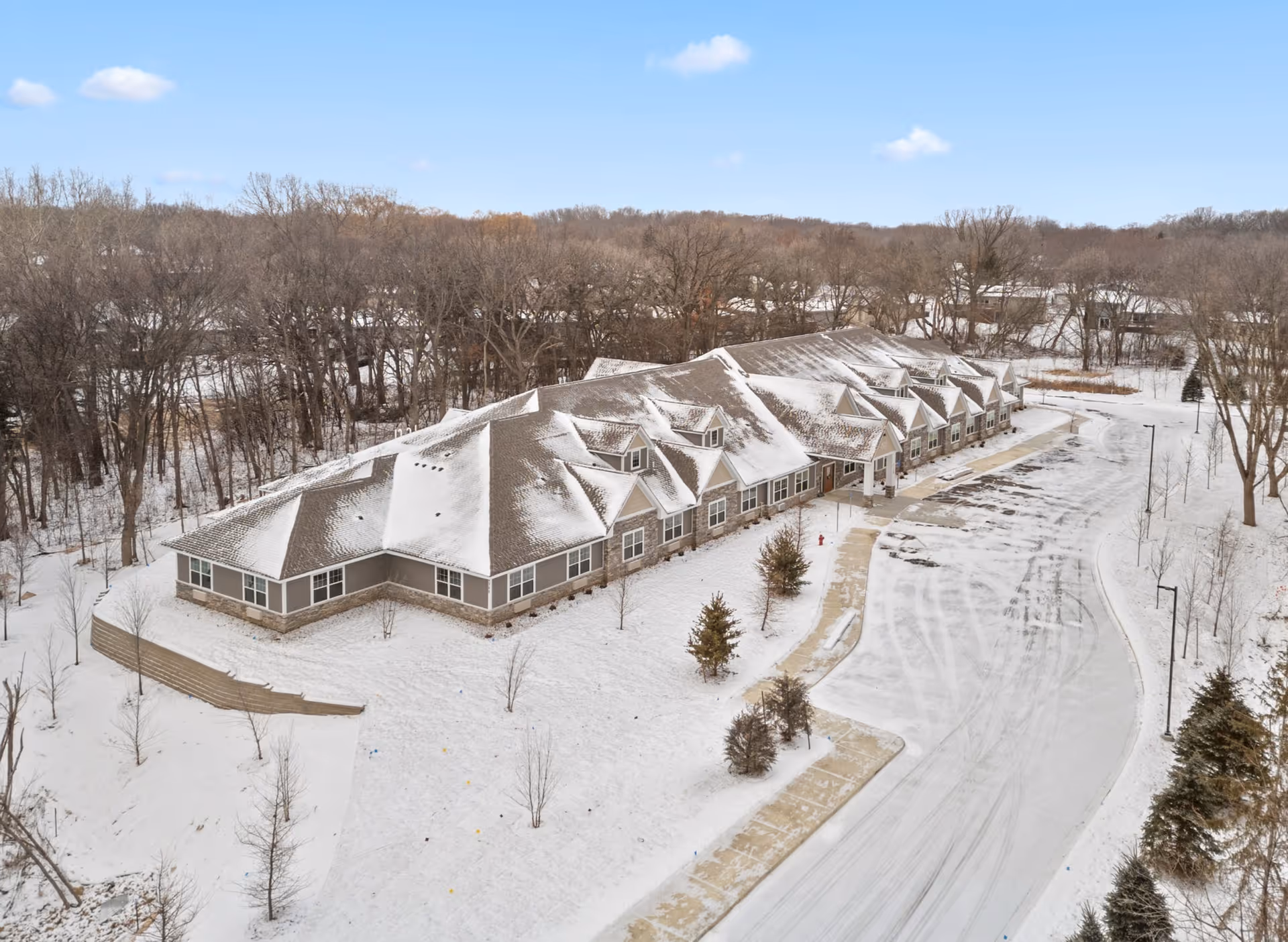 Aerial view of a single-story building and snow-covered parking lot surrounded by trees.
