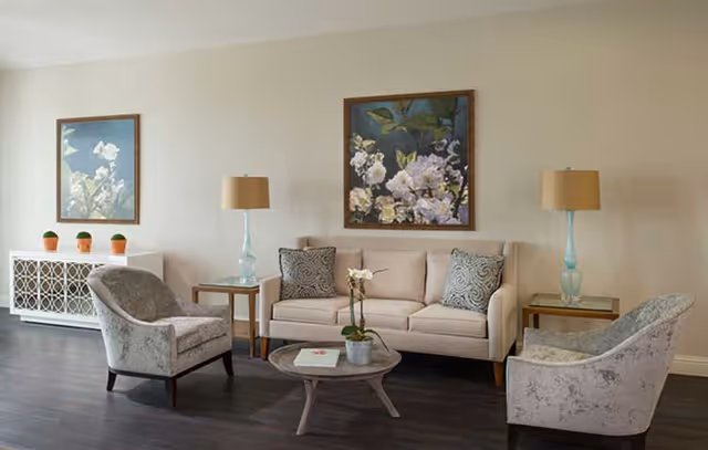 A cozy living room with a beige sofa adorned with two patterned pillows, two light gray armchairs, a round wooden coffee table with a small potted plant and a book, two side tables each holding a glass lamp with a beige shade, a white cabinet with decorative lattice doors topped with three small potted plants, and two framed floral paintings on the wall.