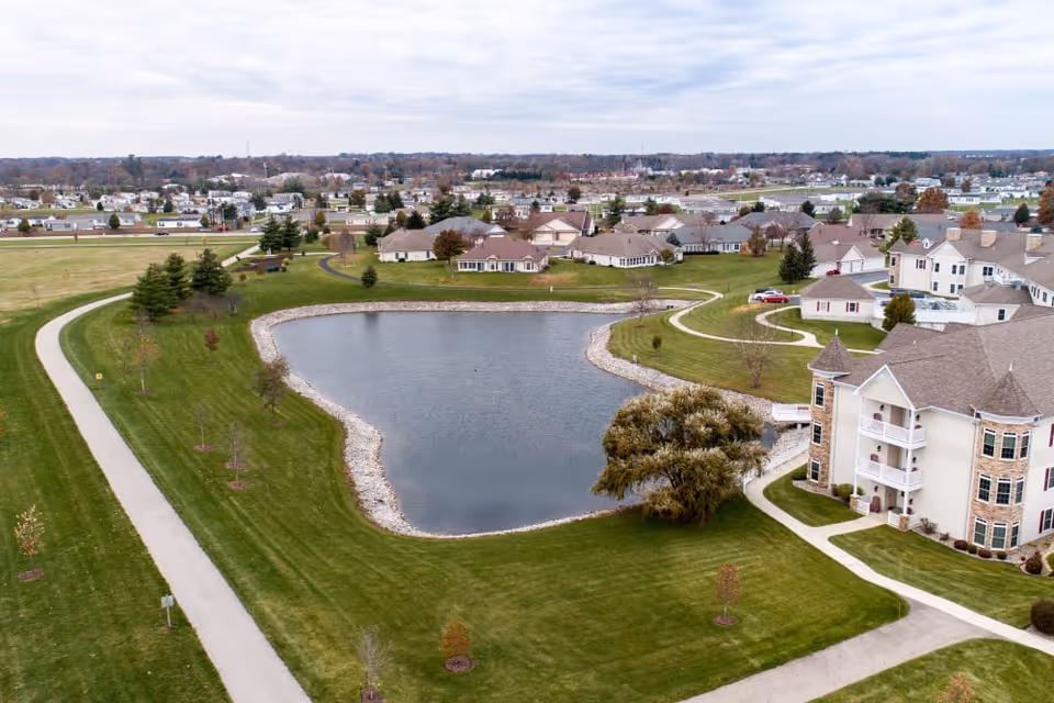 Aerial view of a senior living campus with a central pond, surrounding walkways, lawns, and multi-story residential buildings.