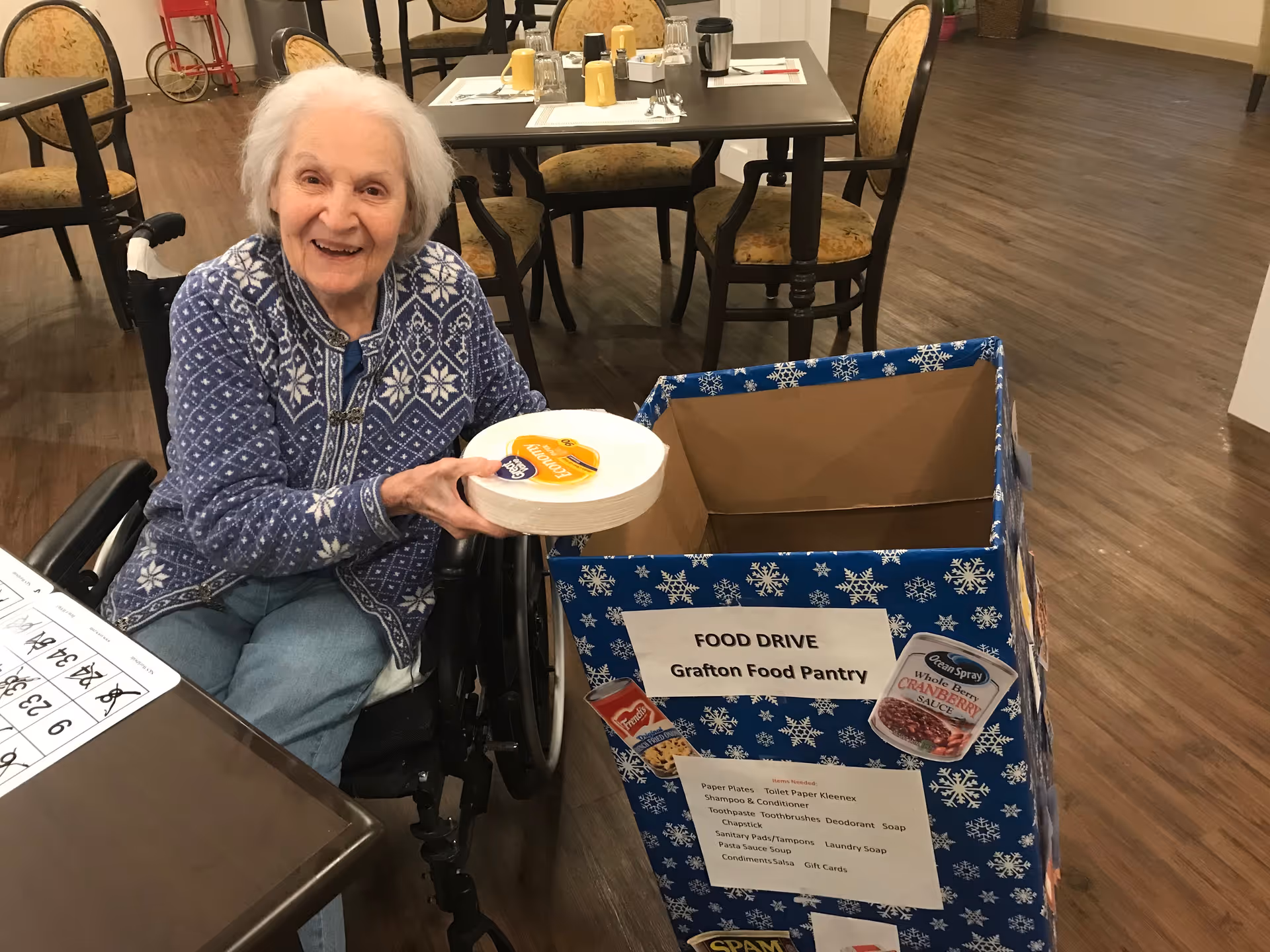 An elderly woman in a wheelchair smiling and holding a stack of paper plates next to a large blue donation box decorated with snowflakes labeled 'FOOD DRIVE Grafton Food Pantry' inside a dining area with tables and chairs.