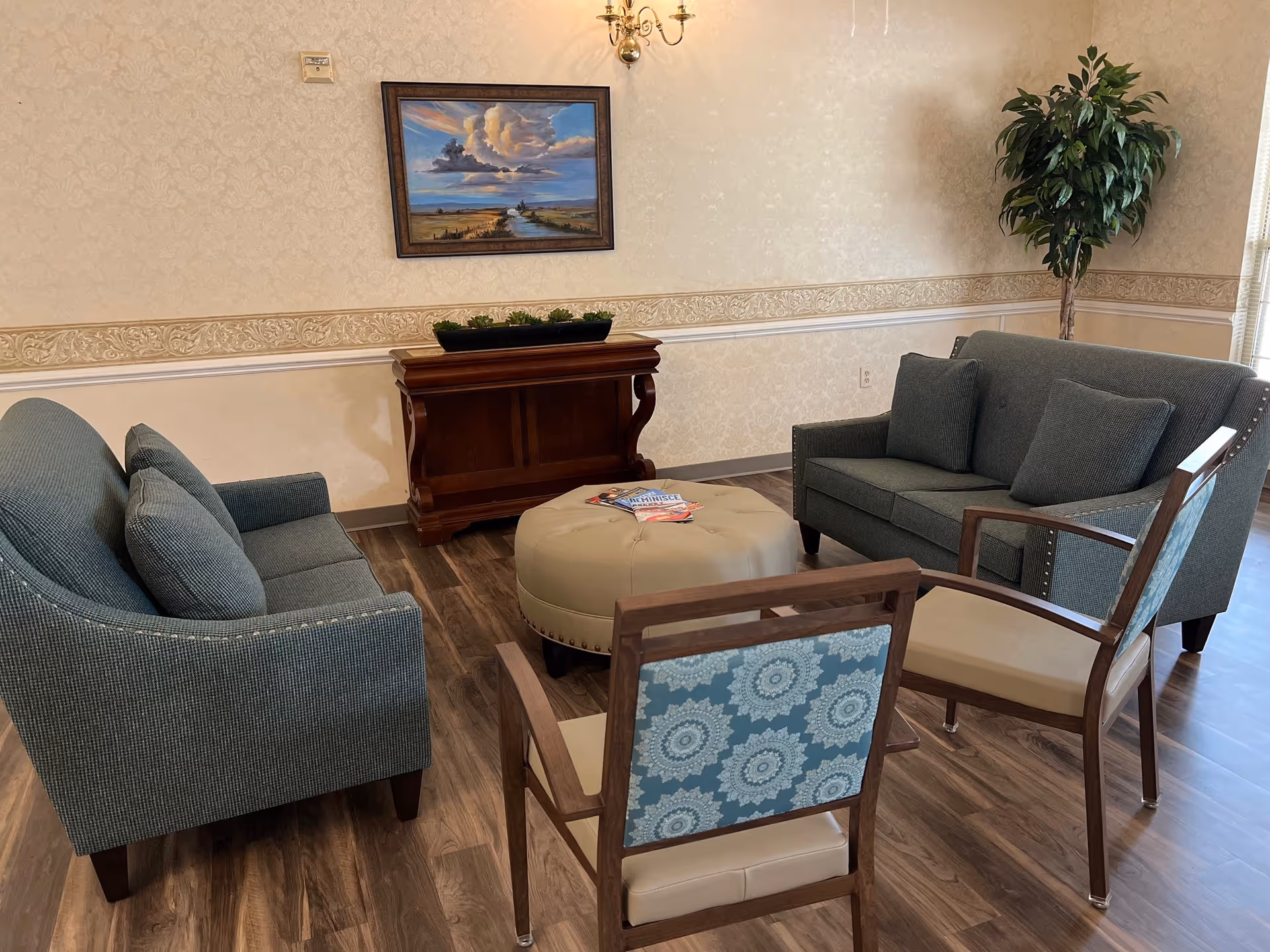 A cozy lounge with two upholstered sofas, two chairs and a round ottoman arranged on wood flooring beneath a framed painting and wall sconce.