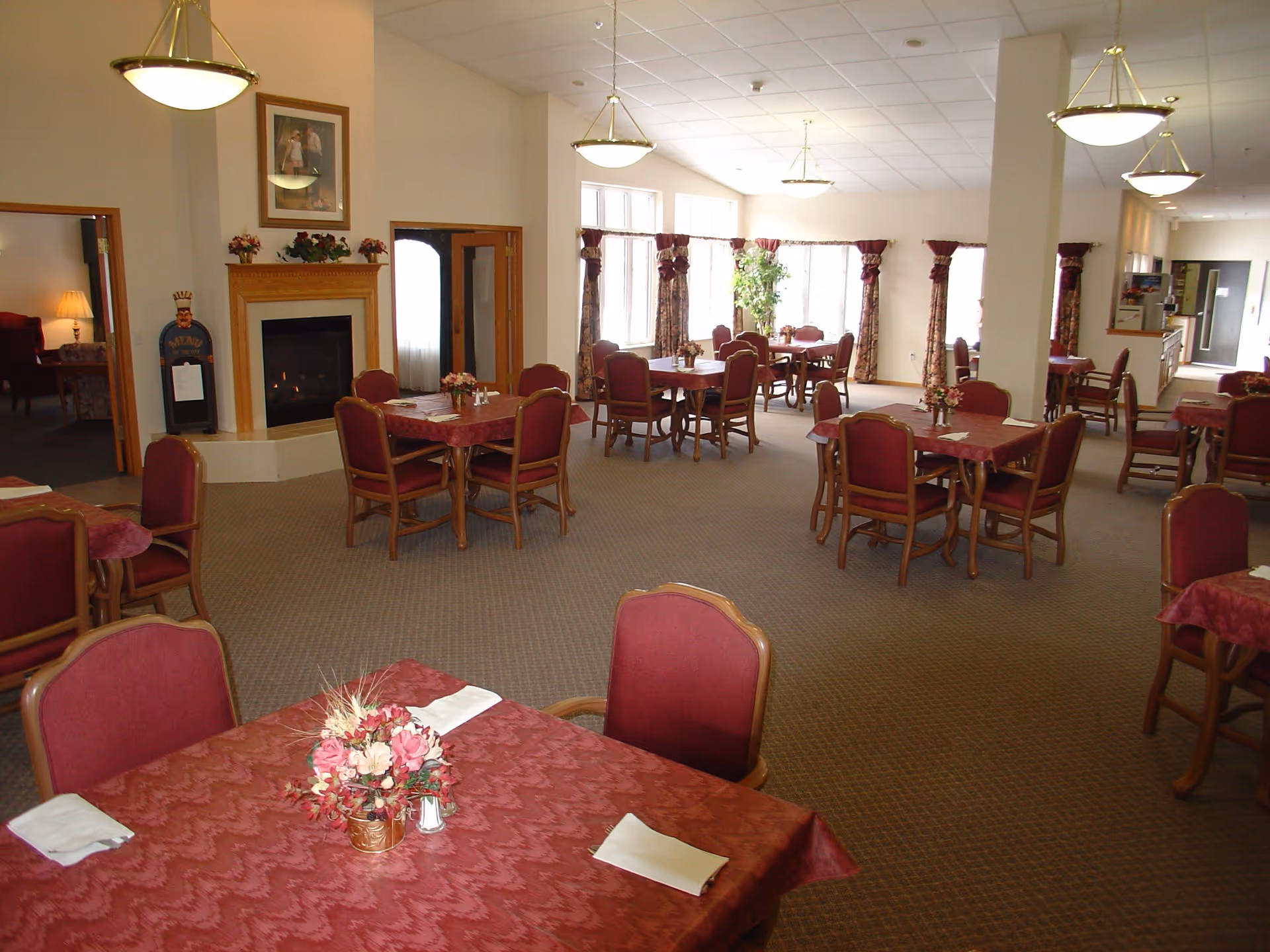 A spacious dining room with multiple tables covered in red tablecloths and surrounded by red cushioned chairs. Each table has a small floral centerpiece and napkins. The room has large windows with floral curtains, a fireplace with a wooden mantel, and warm overhead lighting.