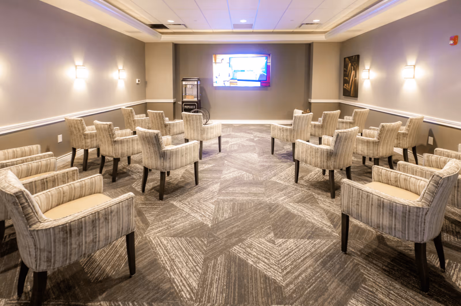 A small theater or media room with multiple beige upholstered armchairs arranged in rows facing a wall-mounted flat screen TV. There is a popcorn machine next to the TV on the left side. The room has patterned carpet flooring, neutral-colored walls, and wall-mounted lights.
