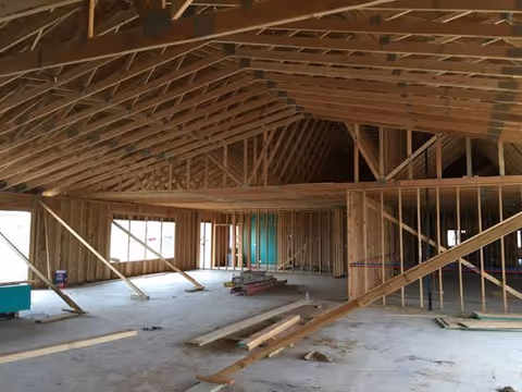 Interior view of a building under construction showing wooden framing and roof trusses with an unfinished concrete floor and construction materials scattered around.