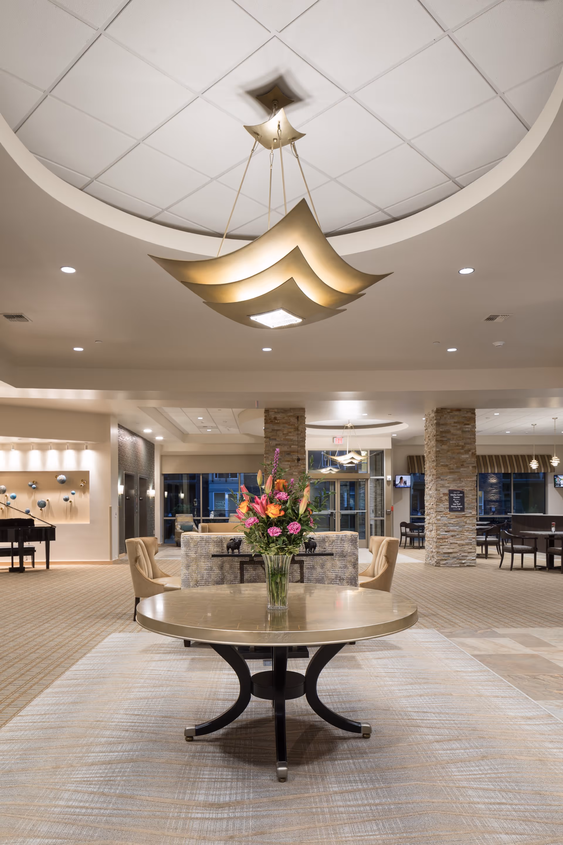 A spacious and well-lit senior living facility lobby with a round table in the center holding a vase of colorful flowers. The ceiling features a modern, artistic light fixture. In the background, there are comfortable seating areas, stone pillars, and large windows.