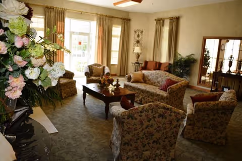 Comfortable living room with floral-upholstered sofas and armchairs arranged around a wooden coffee table, large windows with drapes, and floral arrangements.