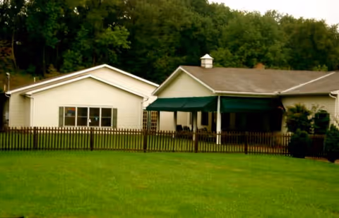 Exterior view of a single-story building with white siding and a green awning over a porch area, surrounded by a wooden fence and a well-maintained grassy lawn, with trees in the background.