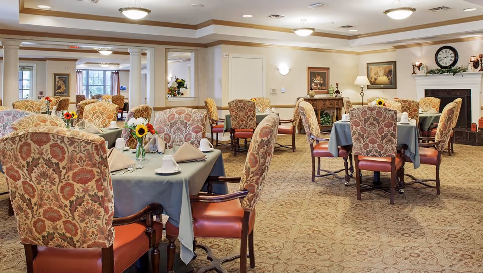 A spacious dining room with multiple tables covered in blue tablecloths, each set with white cups, napkins, and silverware. The chairs have a floral patterned upholstery with wooden arms and legs. The room features beige walls, carpeted floors with a patterned design, and decorative elements including framed paintings, a clock above a white fireplace, and floral centerpieces on the tables.