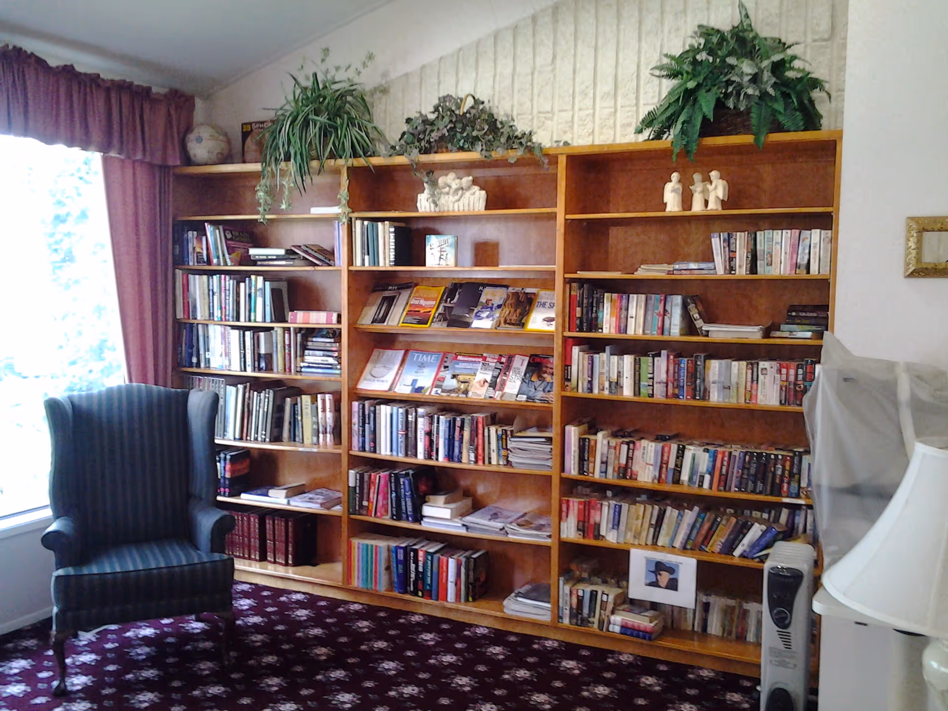 A cozy reading area featuring a large wooden bookshelf filled with books and magazines, a striped upholstered armchair near a window with maroon curtains, and a white lamp on the right side. There are also some decorative plants on top of the bookshelf and small figurines on the shelves.