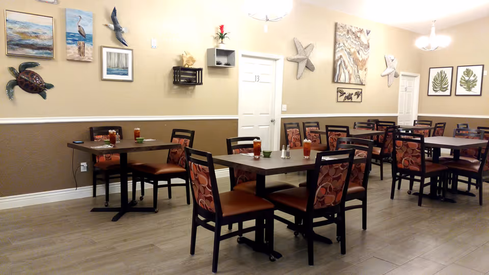 Bright dining room with multiple square tables and patterned chairs, wall art, and glasses of iced tea on the tables.