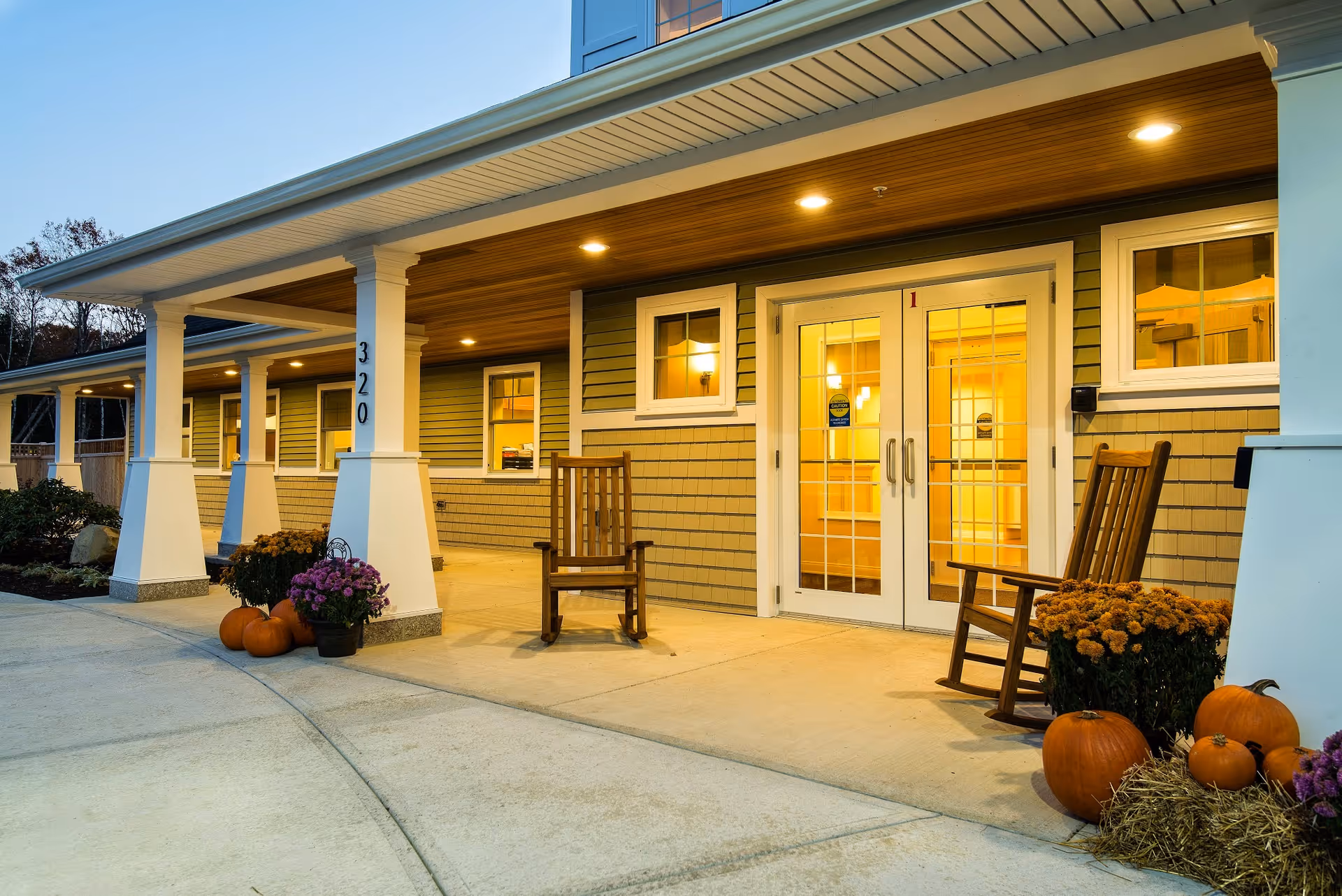 Exterior view of the entrance to Avita of Stroudwater facility at dusk, featuring a covered porch with white pillars, two wooden rocking chairs, pumpkins, potted flowers, and warm lighting inside visible through glass double doors.
