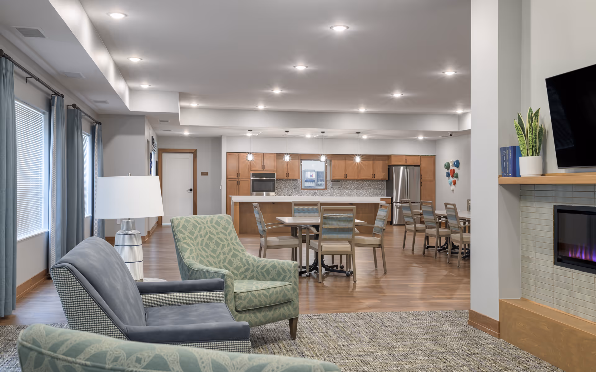 A spacious and modern senior living common area featuring comfortable seating with two armchairs and a sofa, a large open kitchen with wooden cabinets, stainless steel appliances, and multiple dining tables with chairs. The room has wooden flooring, recessed ceiling lights, large windows with curtains, and a fireplace with a mounted TV above it.