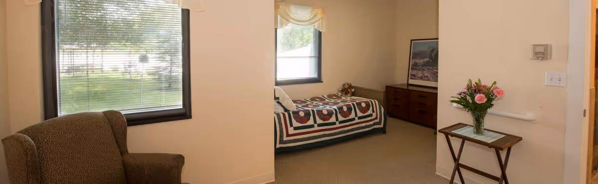 A cozy room with a single bed covered by a colorful quilt, a teddy bear on the bed, a wooden dresser with a framed picture above it, a small table with a vase of flowers, and a comfortable armchair near a window with blinds showing a green outdoor area.