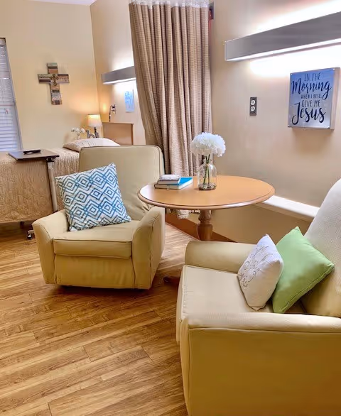 A cozy nursing facility room with two beige armchairs, each with decorative pillows, positioned around a small round wooden table with a vase of white flowers and a book. In the background, there is a bed with a beige quilt, a bedside table with a lamp, a cross hanging on the wall, and a window with closed blinds. A wall decoration with the text 'In the Morning Give Me Jesus' is also visible.