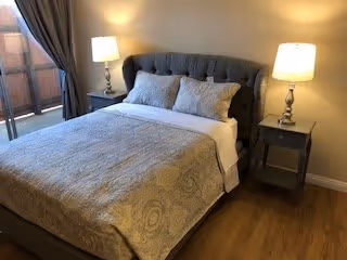 A cozy bedroom with a neatly made bed featuring a tufted gray headboard, three decorative pillows, and a patterned bedspread. There are two matching bedside tables, each with a lamp emitting warm light. A window with curtains is partially visible on the left side.