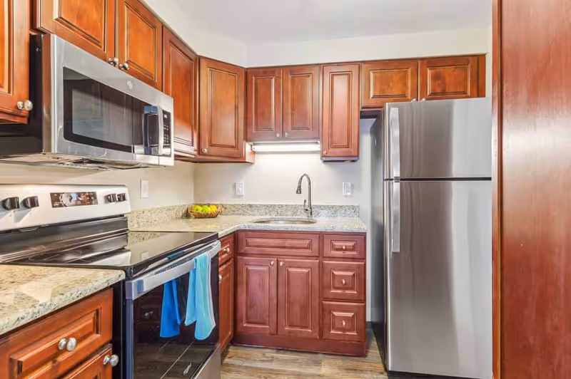 A modern kitchen with wooden cabinets, granite countertops, a stainless steel refrigerator, a microwave, an electric stove, and a sink with a faucet. Two blue towels hang on the oven handle.