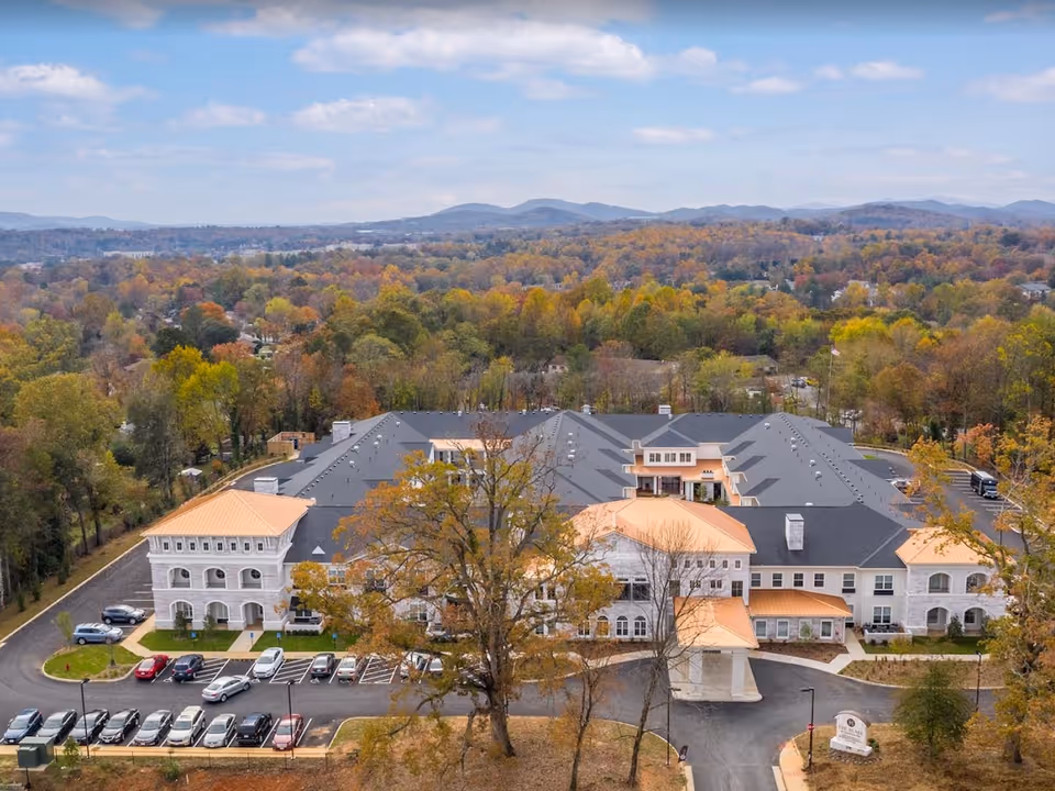 Aerial view of The Blake at Charlottesville senior living facility surrounded by trees with autumn foliage and distant hills under a partly cloudy sky. The building has a large parking lot with multiple cars parked and a driveway leading to the entrance.