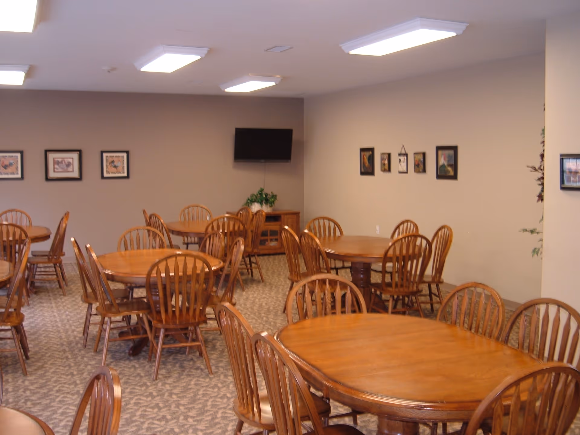 A dining room with multiple wooden round and oval tables surrounded by wooden chairs. The room has beige walls decorated with framed pictures, a carpeted floor, and a flat-screen TV mounted on the wall above a wooden cabinet with a plant on top.