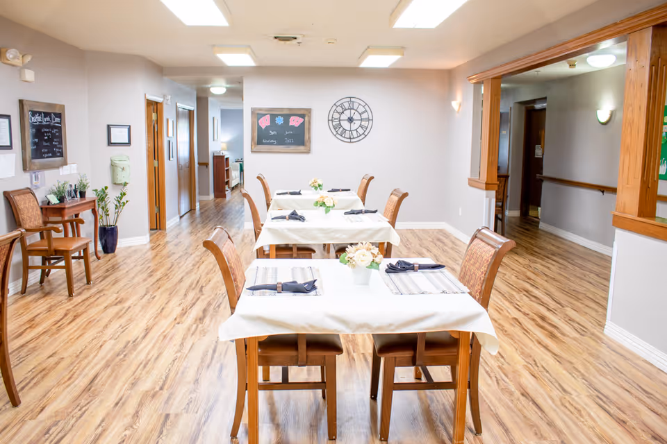 A bright dining room with wooden floors and three tables covered with white tablecloths, each set with placemats, napkins, and small flower arrangements. The room has wooden chairs, a large wall clock, and framed chalkboards on the walls. There is a hallway visible in the background and wooden trim around an open pass-through window on the right.