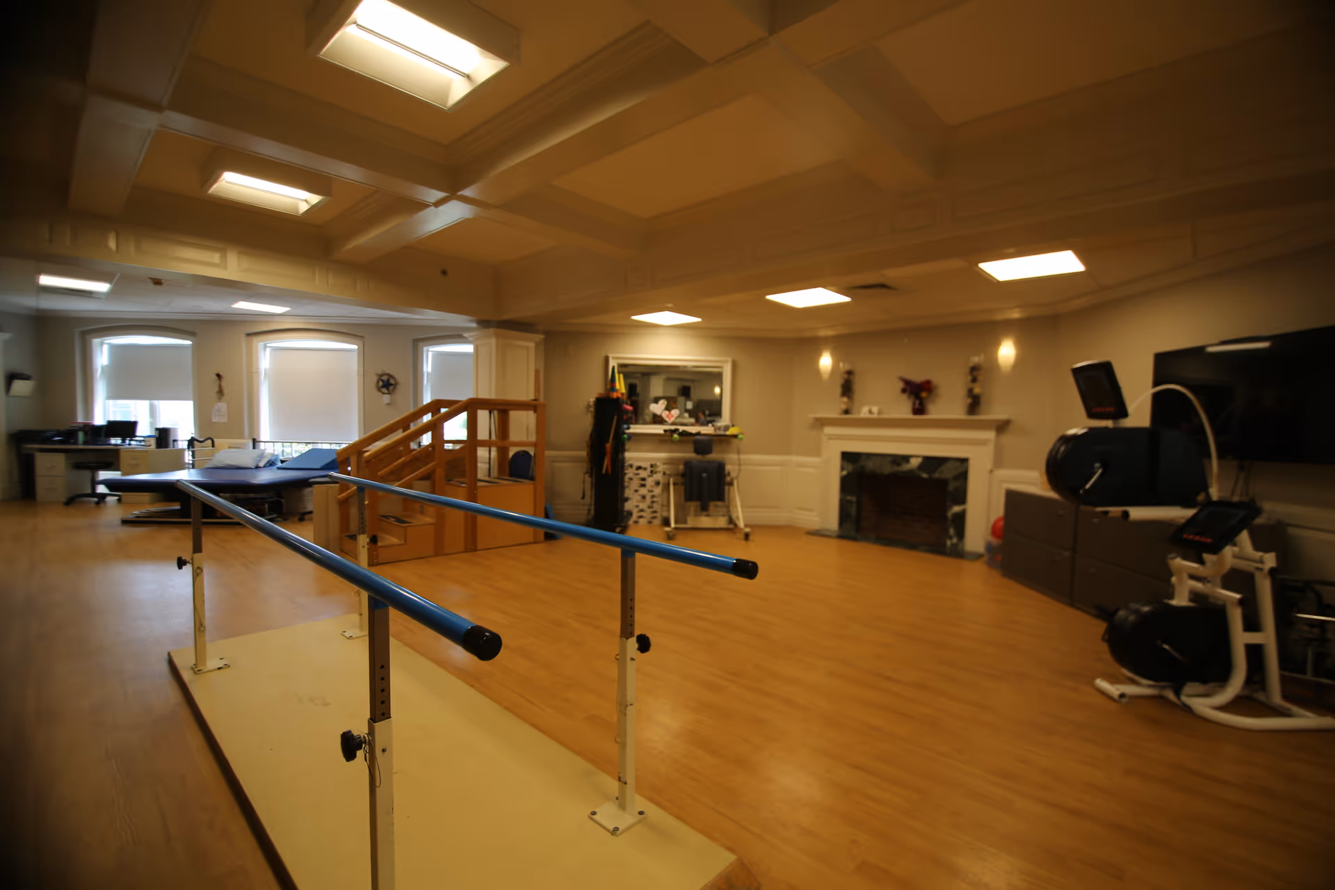 A spacious rehabilitation or physical therapy room with wooden flooring, parallel bars for walking exercises, a small staircase with handrails, exercise equipment, a desk area with chairs near windows, and a fireplace on the far wall under bright ceiling lights.